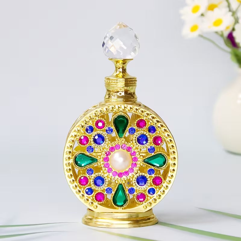 A decorative gold perfume bottle with a clear faceted crystal stopper, adorned with colorful faux gemstones in green, blue, pink, and a central white pearl, set against a light gray background with a blurred white flower vase in the corner.